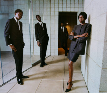 A group of four individuals is stylishly dressed in formal attire, featuring suits and elegant shoes. They stand against a backdrop of concrete walls and polished tiles. Their expressions convey confidence and sophistication as they pose strategically around a glass-paneled area. The composition is balanced, highlighting both their attire and the modern setting.