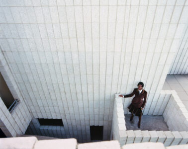 A person stands on a gray tiled rooftop terrace, wearing a dark coat. The perspective showcases tall, textured concrete walls composed of rectangular blocks, creating a geometric pattern. The setting appears open and expansive, with a modern architectural design. The overall atmosphere is serene yet striking, characterized by contrasting shades of gray and brown.