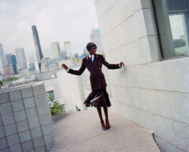 A stylish individual in a dark, tailored suit joyfully balances on a narrow outdoor path, bordered by modern architectural elements. The city skyline, with various skyscrapers under a partially cloudy sky, forms the backdrop. The person's playful stance and flowing skirt interaction with the urban environment create a dynamic, fashion-forward scene, blending elegance with contemporary urban life.