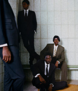 Four individuals in formal black and brown suits are positioned in a tiled room. One stands against the wall, another sits on a bench, and two are seated on the floor. The atmosphere appears contemplative, with each person gazing in different directions. The setting conveys a sense of seriousness and introspection, with muted colors and a quiet, introspective ambiance.