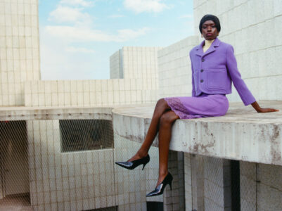 A person sits confidently on the edge of a concrete platform, dressed in a chic lavender outfit consisting of a jacket and skirt, complemented by black heeled shoes. The geometric architecture of white, block-like structures surrounds them, under a partly cloudy sky, creating a harmonious blend of fashion and modern architectural elements.