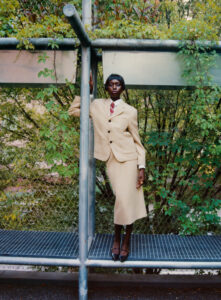 A person wearing a beige suit and tie stands confidently on a metal platform. The setting features a background of lush green foliage and metal structures. The attire includes a tailored jacket and skirt, complemented by heeled shoes. The scene conveys a blend of elegance and urban aesthetic, highlighting a contrast between fashion and natural elements.