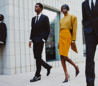 Two individuals walk confidently on an urban sidewalk, surrounded by modern architecture with textured white walls. The person on the left wears a sharp black suit and glossy shoes, while the individual on the right dons a striking mustard ensemble with sleek high heels. Their professional and fashionable appearances exude sophistication and elegance in the city setting.