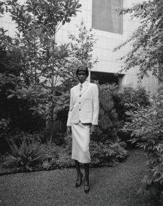 A figure dressed in a tailored suit and skirt stands poised among lush foliage. The setting appears to be a garden or courtyard with a gravel pathway. The towering plants and trees provide a natural backdrop, complementing the modern architecture of a building in the background. The atmosphere feels serene and sophisticated, blending nature with style.