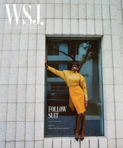 A person stands confidently in front of a large window, framed by a grid-patterned stone facade. Dressed in a mustard yellow skirt and top, they wear high heels, one arm resting on the frame. Reflections show a tree and building, adding depth. The pose is stylish and poised, evoking a sense of elegance and modernity.