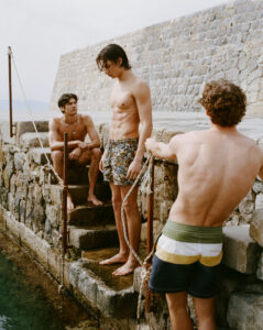 Three men in swimwear gather on stone steps beside calm water, framed by a weathered wall. One stands with head slightly bowed, another sits watching him, while a third leans forward gripping a rope. Their postures feel quiet and reflective. Warm, natural light enhances skin tones and textures, creating a relaxed, intimate moment in a coastal setting.