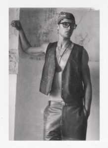 A person stands confidently against a textured wall, wearing a leather vest and loose-fitting pants. Their head is adorned with a patterned bandana, and they sport large glasses, adding a vintage touch. Accessories include a necklace and a belt, contributing to the stylish ensemble. The setting conveys a rustic, timeless aesthetic, enhancing the individual's fashion-forward appearance.