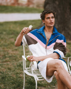 A person with short hair sits relaxed on a white metal chair outdoors. They wear a colorful zip-up jacket with blue, pink, brown, and white stripes, paired with white shorts. Holding a slice of fruit, they appear contemplative. The setting is grassy, with a blurred background hinting at trees or park scenery, creating a serene atmosphere.