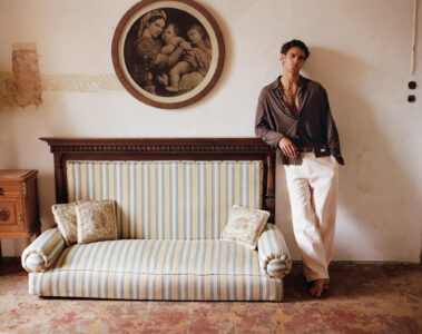 A person leans casually against a rustic wall, combining elegance and relaxation. Their attire blends a dark, open-collared shirt with light pants. Behind them, a vintage, striped sofa with decorative cushions is positioned beneath a circular framed artwork depicting a classical scene. The floor has a worn, earthy tone, and the ambiance is one of understated sophistication.