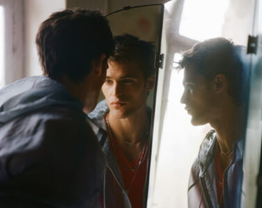 A young man gazes into a mirror, creating a reflective effect. He wears layered clothing with a chain around his neck. The soft lighting casts gentle shadows, highlighting his contemplative expression. The scene is set in a softly lit room with a window, adding a serene ambiance, and the mirror angles capture different facets of his face.