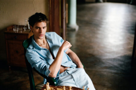 A person sits in a relaxed pose on a wooden chair, wearing a light blue outfit with an unbuttoned shirt, revealing a hint of jewelry. The background features a dimly lit room with hardwood flooring and a wooden cabinet holding glassware. The atmosphere is serene and contemplative, with a chessboard hinting at a quiet leisure activity.