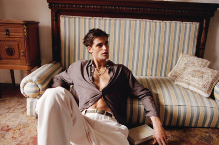 A person lounges comfortably against a striped sofa with a vintage wooden backdrop. They wear a partially unbuttoned dark shirt and light trousers. A book rests beside them on a patterned floor, suggesting leisure and relaxation. Ornate cushions add an elegant touch to the scene, complementing the overall warm and sophisticated ambiance of the space.