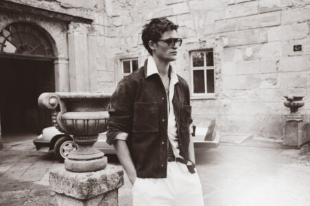 A stylish individual stands in a vintage courtyard, wearing sunglasses and a casual outfit featuring a dark jacket over a light shirt, paired with white pants. The scene features an ornate stone planter and a classic car in the background. The architecture is rustic, with textured stone walls and arched windows, evoking an old-world charm.