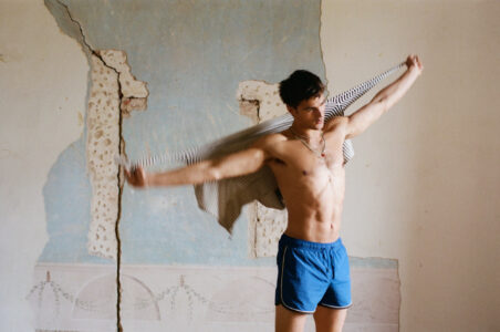 A shirtless man stands against a textured, cracked wall with an old mural. He wears blue shorts and holds a striped shirt, seemingly putting it on or taking it off. The background features faded blue paint and exposed brick, adding a feeling of rustic decay. The scene captures a moment of casual movement and understated elegance.