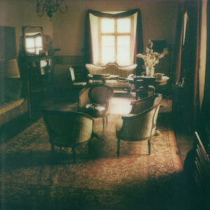 A vintage-style living room exudes warmth through soft, natural light filtering from a wide window with draped curtains. Elegant, striped chairs and a sofa encircle a dark wooden table adorned with delicate flowers. An ornate chandelier hangs from the ceiling, highlighting the intricate patterns of a large, plush rug that covers the wooden floor. A cozy, serene ambiance prevails.