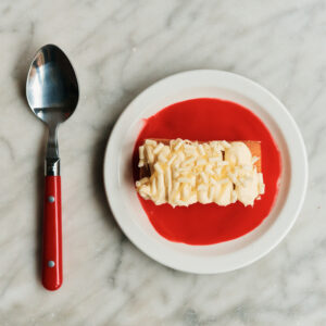 A creamy dessert sits on a small white plate with vibrant red sauce. The dessert features a smooth texture topped with grated white chocolate. Adjacent to the plate is a metal spoon with a red handle, all set upon a grey marbled surface. The arrangement creates a striking contrast between the creamy hues and vivid colors.