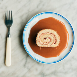 A marble surface holds a white plate with blue rim containing a dessert. The dessert is a slice of rolled cake with a swirl pattern, sitting atop thick caramel sauce. Beside the plate, a fork with a dark handle and lighter grip rests on the surface. The presentation is simple yet appetizing.
