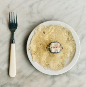 A table setting includes a small white plate with translucent, golden jelly-like substance and a single piece of fish placed centrally. A fork with a black handle sits to the left. The table surface features a light marble pattern with subtle veining. The overall arrangement is minimalistic, emphasizing the contrast between the dish and the background.