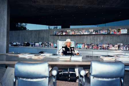 A person sits in a sleek, modern concrete space, surrounded by an array of vibrant photographs on display shelves. The individual wears a wide-brimmed hat and casual attire, seated at a large desk with papers and magazines. The setting features stylish leather chairs and recessed ceiling lights, complementing the contemporary, sophisticated ambiance.