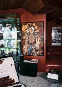 A cozy room features a reddish-brown wall with an artwork depicting a person in a hat and patterned clothing. A glass display case on the left holds assorted framed photos. A cowhide-patterned chair and magazines are nearby. A corner of a red leather couch peeks from the right, alongside a table stacked with books.
