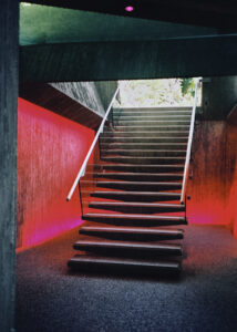 A modern staircase with railings on either side ascends to an upper level, framed by striking red and pink lighting. The walls have a textured, concrete appearance, adding to the contemporary ambiance. A glimpse of bright greenery is visible at the top, offering a contrast to the otherwise industrial and moody atmosphere.