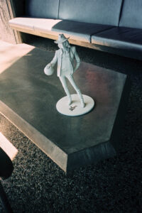 A small figurine of a person wearing a hat and holding a ball stands on a geometric platform. Shadows from the afternoon sun cast across the smooth surface. A modern, minimalist couch sits in the background, suggesting a contemporary interior setting. The floor is covered with textured pebbles, adding contrast to the sleek and polished surfaces.