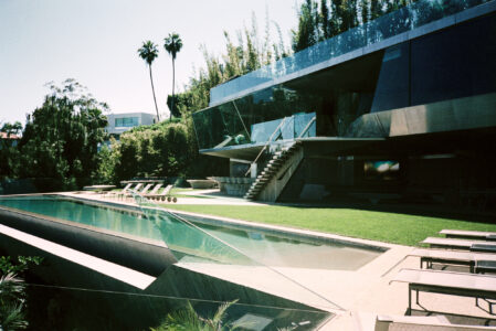 A modern architectural marvel featuring sleek lines and extensive use of glass, surrounded by lush greenery. A sparkling infinity pool stretches alongside the building, bordered by several lounge chairs. The upper level features a terrace with transparent railings, while a staircase connects the two levels. Tall palm trees rise gracefully in the background, completing the serene and luxurious setting.
